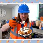 Man in blue hard hat and orange safety vest holds a waffle bowl topped with whipped cream and chocolate sauce in a cafe kitchen; a cartoon construction worker stands on the right.