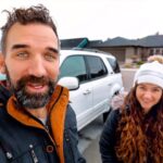 Smiling man and woman take a selfie in a residential driveway, with a white SUV and a blue car in the background.
