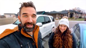 Smiling man and woman take a selfie in a residential driveway, with a white SUV and a blue car in the background.