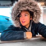 Person in a fur-hooded coat lies on the garage floor under a car, looking up.