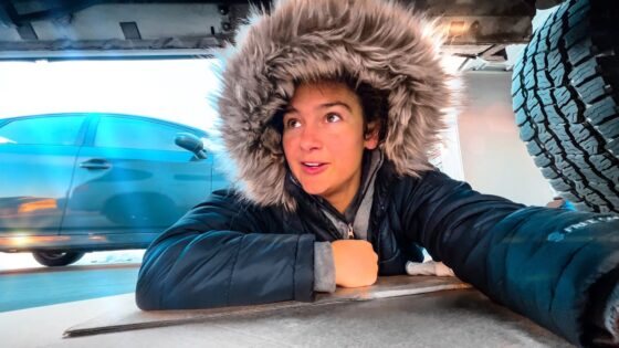 Person in a fur-hooded coat lies on the garage floor under a car, looking up.