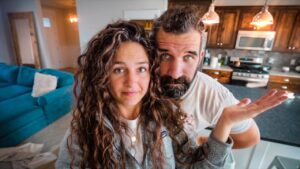 Couple in a kitchen; woman with curly hair in front, man with beard leaning over from behind and gesturing with an open hand.