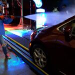 Worker wearing a blue jacket and helmet uses a high-pressure nozzle to spray a car at a car wash bay, colorful lights around.