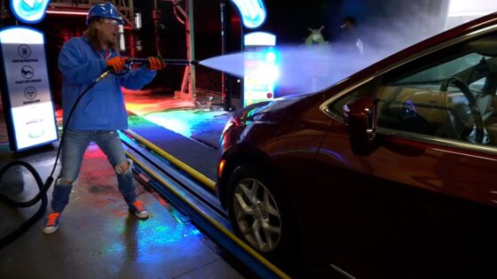 Worker wearing a blue jacket and helmet uses a high-pressure nozzle to spray a car at a car wash bay, colorful lights around.