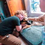 Children kneel and cuddle a dog lying on a blue rug inside a home, comforting it with gentle pets.