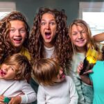 Group of kids with wild hair posing in a bright room, two raising large scissors as if styling hair and others making goofy faces.