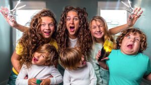 Group of kids with wild hair posing in a bright room, two raising large scissors as if styling hair and others making goofy faces.