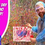 Man in a blue poncho and safety goggles paints a colorful splatter artwork on a small canvas, with a vibrant Valentine’s Day Art Studio backdrop behind him.