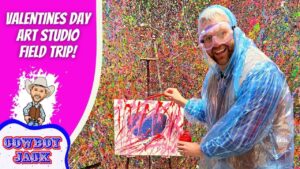 Man in a blue poncho and safety goggles paints a colorful splatter artwork on a small canvas, with a vibrant Valentine’s Day Art Studio backdrop behind him.