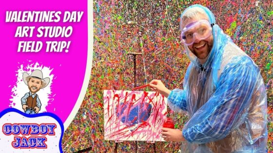 Man in a blue poncho and safety goggles paints a colorful splatter artwork on a small canvas, with a vibrant Valentine’s Day Art Studio backdrop behind him.