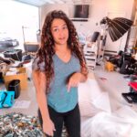 Woman stands in a cluttered garage studio filled with gear, boxes, and a large lighting setup behind her.