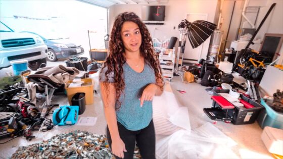 Woman stands in a cluttered garage studio filled with gear, boxes, and a large lighting setup behind her.
