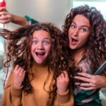 Two women with long curly hair having fun while styling hair; one holds a red curling iron and lifts the other's curls, both smiling in a bright bedroom.