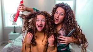 Two women with long curly hair having fun while styling hair; one holds a red curling iron and lifts the other's curls, both smiling in a bright bedroom.