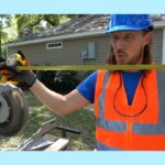 Construction worker in a blue hard hat and orange safety vest holds a level across a miter saw outdoors in front of a house.
