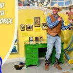Man in a cowboy hat and vest poses beside a Curious George exhibit with a green desk and yellow wall.
