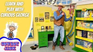 Man in a cowboy hat and vest poses beside a Curious George exhibit with a green desk and yellow wall.