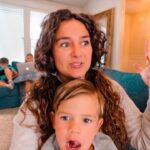 Woman with curly hair on a blue couch raises her hand while a child sits in front; other kids on laptops sit on the sofa in the background.