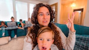 Woman with curly hair on a blue couch raises her hand while a child sits in front; other kids on laptops sit on the sofa in the background.