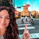 Woman with long hair in foreground points toward a city square with cyclists crossing a crosswalk and a domed capitol building in warm sunset light behind them.
