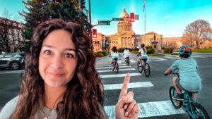 Woman with long hair in foreground points toward a city square with cyclists crossing a crosswalk and a domed capitol building in warm sunset light behind them.