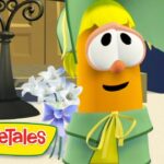 Orange VeggieTales character with large eyes and yellow bow, wearing green outfit, smiling beside a bouquet of white flowers on a stage.
