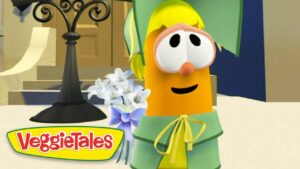 Orange VeggieTales character with large eyes and yellow bow, wearing green outfit, smiling beside a bouquet of white flowers on a stage.