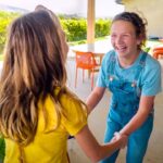 Two women laugh and hold hands on a sunny patio, greeting each other as colorful chairs and greenery frame the scene.