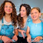 Three people in a home: a girl in denim overalls and a white shirt on the left, a woman in the middle talking, and a girl in a blue striped shirt on the right holding papers.