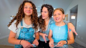 Three girls in a bright living room posing for a photo: one with curly hair in denim overalls, another with a gray hoodie, and the third in a blue striped shirt, all holding papers and smiling.