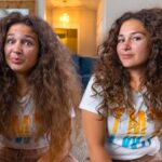 Two young women with long curly hair sit on the living room floor, smiling at the camera in a cozy home setting.