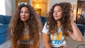 Two young women with long curly hair sit on the living room floor, smiling at the camera in a cozy home setting.