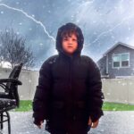Child in a black puffy coat stands outside in falling snow as bright lightning streaks across a dark, cloudy sky behind him.
