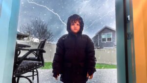 Child in a black hooded winter coat stands in a doorway outside during a snowstorm, with lightning in the cloudy sky behind a fenced yard and patio chair.