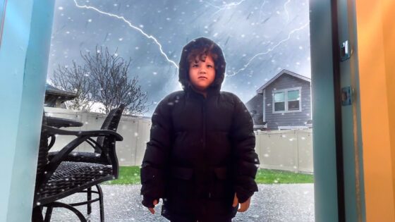 Child in a black hooded winter coat stands in a doorway outside during a snowstorm, with lightning in the cloudy sky behind a fenced yard and patio chair.