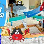 Man in a blue plaid shirt and brown vest sits in a carved wooden pirate ship sandbox with a blue canopy and cartoon decorations for kids activity.