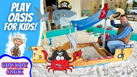 Man in a blue plaid shirt and brown vest sits in a carved wooden pirate ship sandbox with a blue canopy and cartoon decorations for kids activity.