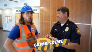Construction worker in an orange safety vest and blue helmet talks with a police officer in a courthouse lobby; a 'CRIME SCENE' overlay is across the image.