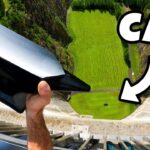 Person grips a large black and silver curved object over a dam, with a green field below and a bold 'CAR' graphic on the right.