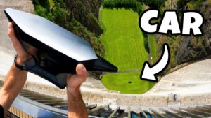 Person grips a large black and silver curved object over a dam, with a green field below and a bold 'CAR' graphic on the right.