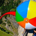 Person in a black shirt holds a giant multicolored beach ball near a cliff edge on a roadway, suggesting a high-risk stunt.