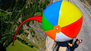 Person in a black shirt holds a giant multicolored beach ball near a cliff edge on a roadway, suggesting a high-risk stunt.