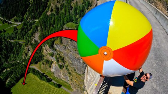 Person in a black shirt holds a giant multicolored beach ball near a cliff edge on a roadway, suggesting a high-risk stunt.