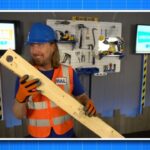 Man in a blue hard hat and orange safety vest holds a long wooden plank in a tool-filled workshop, smiling at the camera.