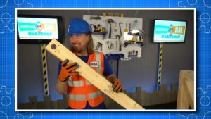 Man in a blue hard hat and orange safety vest holds a long wooden plank in a tool-filled workshop, smiling at the camera.
