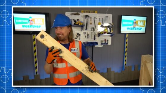 Man in a blue hard hat and orange safety vest holds a long wooden plank in a tool-filled workshop, smiling at the camera.