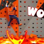 Climber in a safety vest scales an indoor orange-gray climbing wall, with a fiery lava graphic at the bottom and a big WOW! caption overlay.