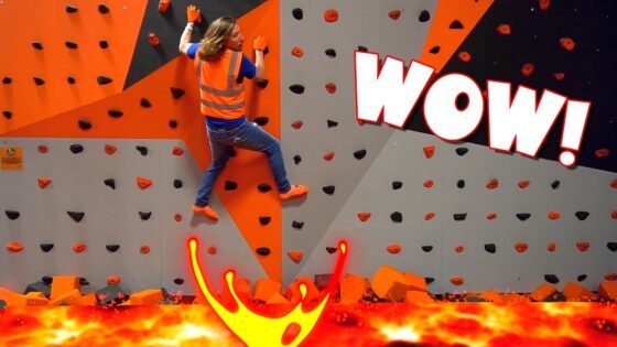 Climber in a safety vest scales an indoor orange-gray climbing wall, with a fiery lava graphic at the bottom and a big WOW! caption overlay.
