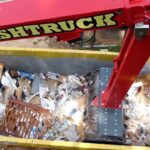 Red Smash Truck arm dumping a yellow dumpster filled with paper and cardboard waste, visible trash and debris inside the bin.