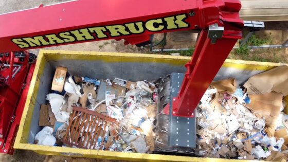 Red Smash Truck arm dumping a yellow dumpster filled with paper and cardboard waste, visible trash and debris inside the bin.